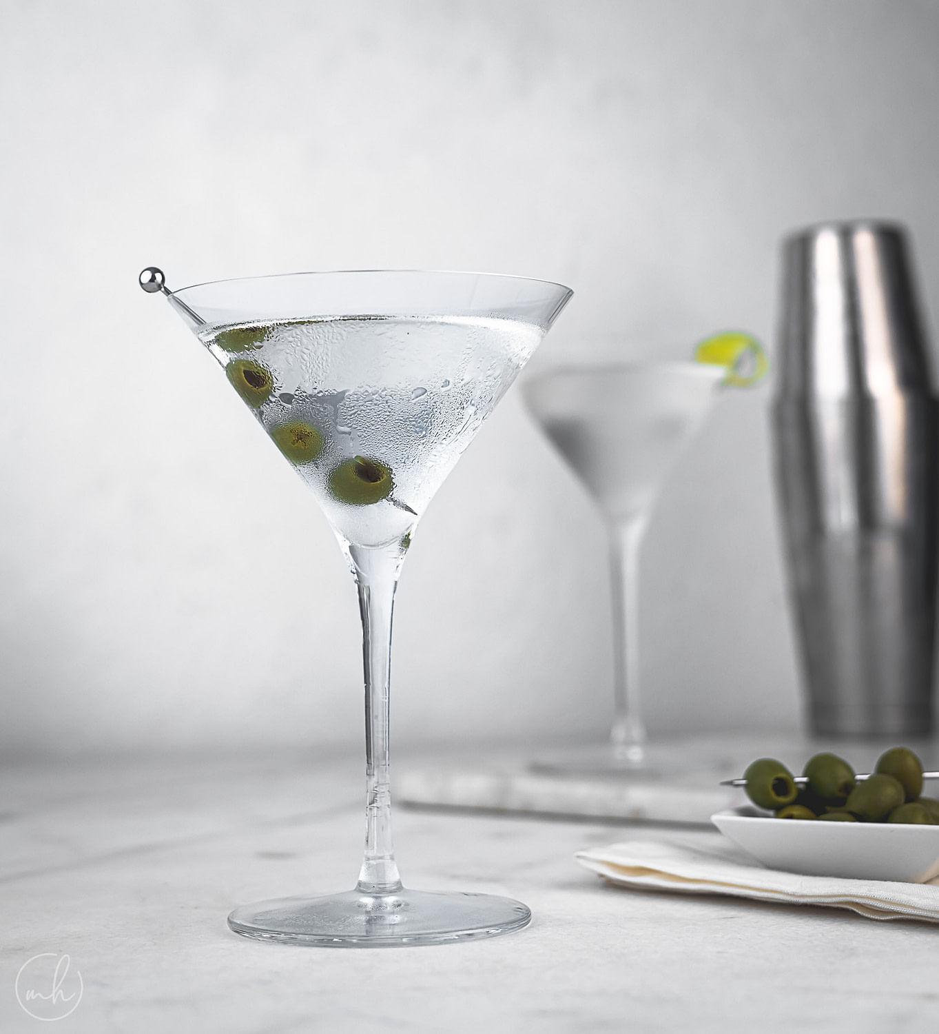 A martini cocktail glass with olives as garnish. In the backgroun, another martini glass, a bowl of olives and a shaker is placed.