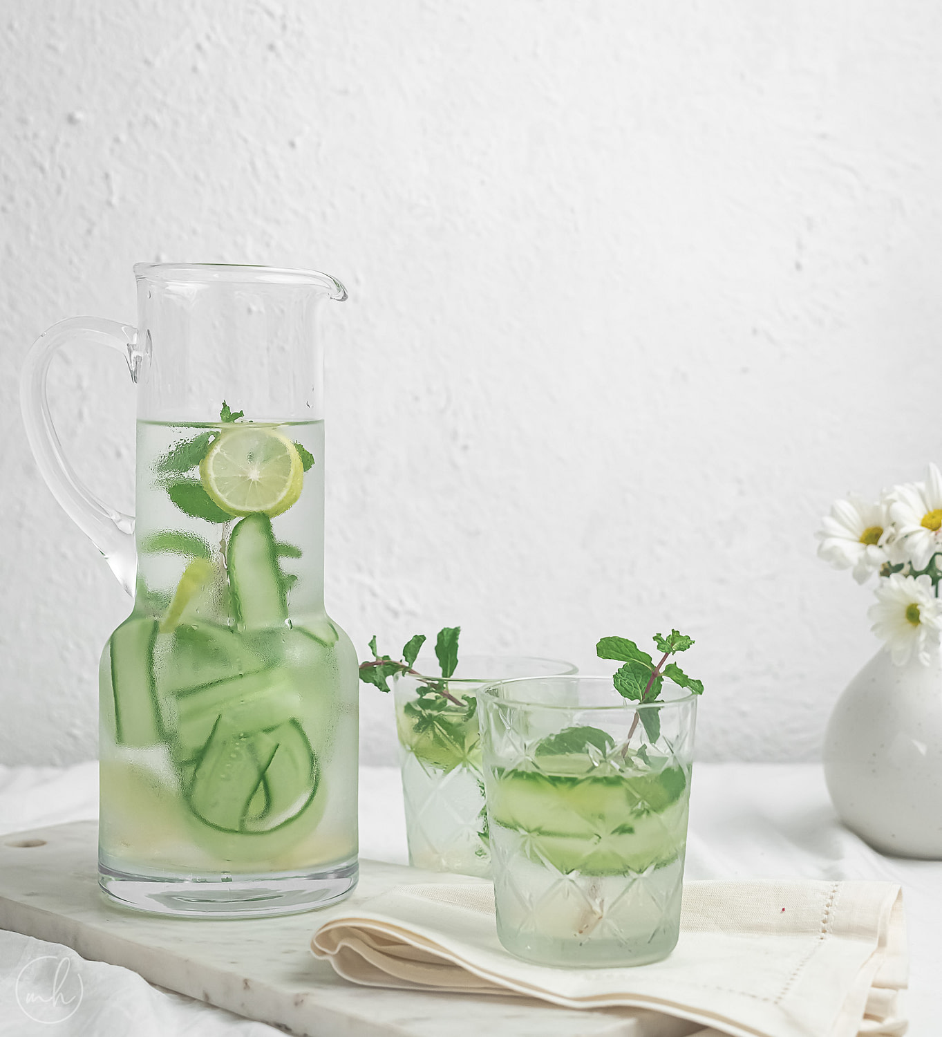 Two glasses of cucumber basil lemonade with cucumber slices as garnish. A potted vase with white flowers, cucumber peels, sliced lemon and a half-filled jar of lemonade is in the background.
