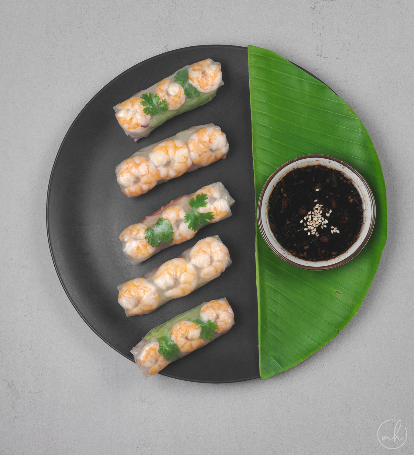 Shrimp summer roll placed on a black plate next to a banana leaf with a soy dipping sauce