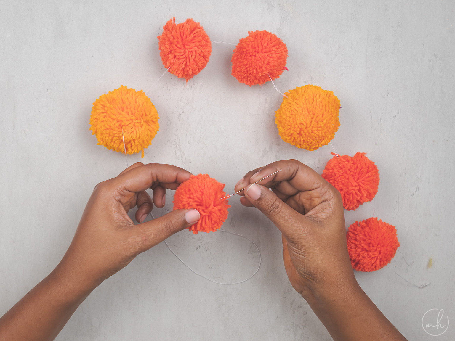 Two hands running a threaded needle through red-yellow pompoms