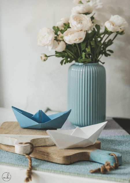 Kagazi: A blue colour paper boat, a white colour paper boat, flowers in a vase against a white backdrop