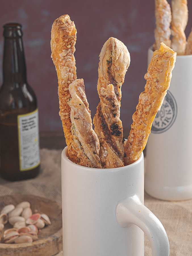Sundried tomatoes and olive puff pastry cheese straws placed in a cup with beer bottle behind