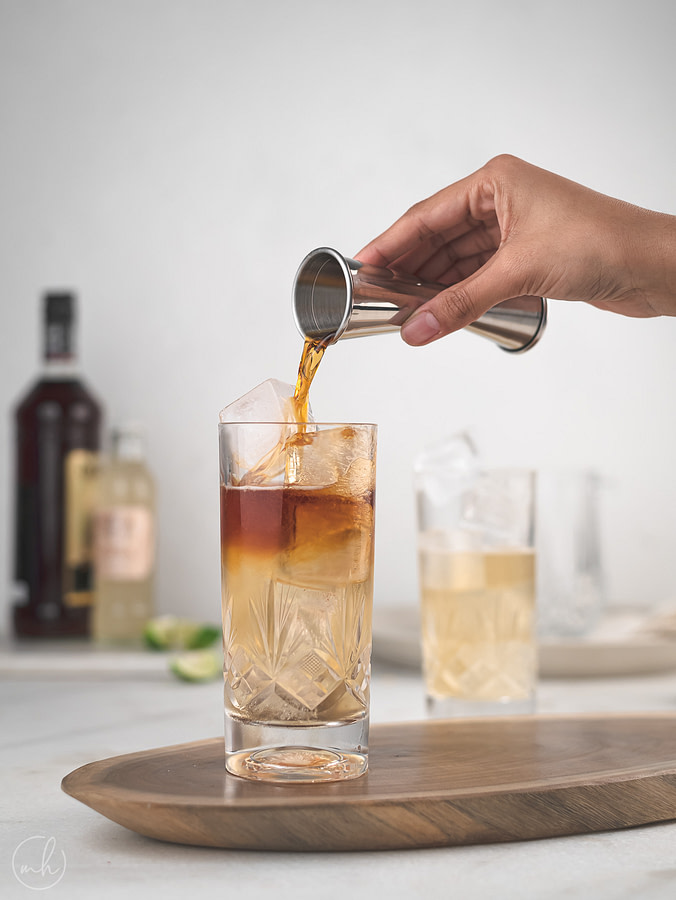 A hand pouring a dark & stormy into a nearly full glass with ice, using a steel jigger. Another glass half full with a liquid behind. Next to it are two bottles - one black and one pink.