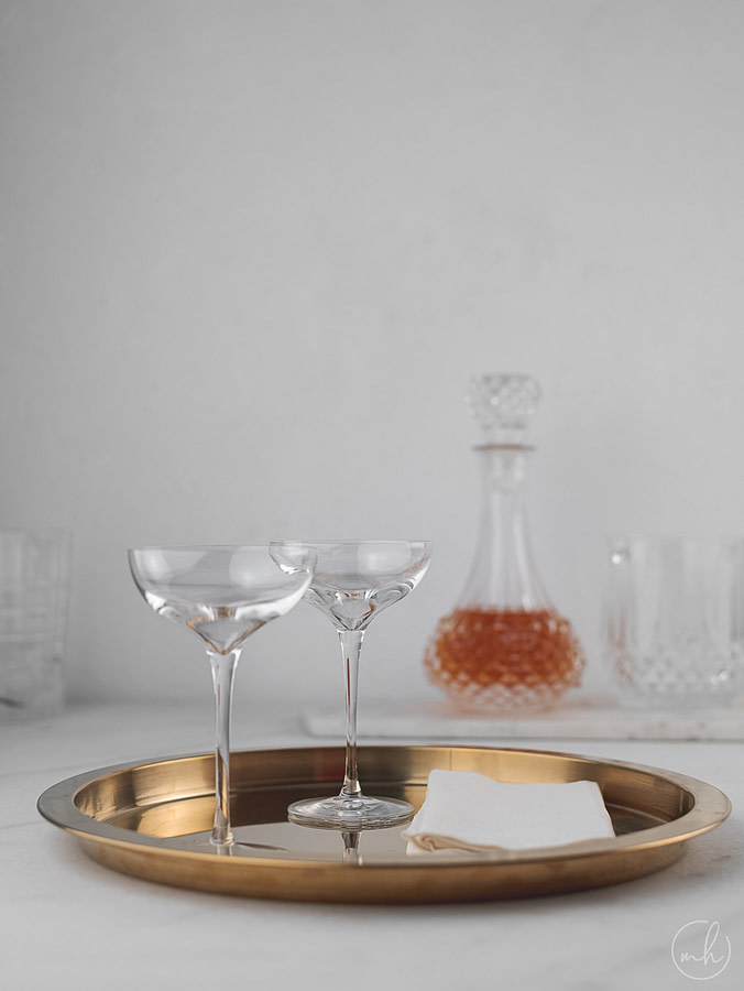 Two empty cocktail glasses placed on a copper tray, with a cloth napkin. In the background, a half-filled decanter of vermouth is placed.