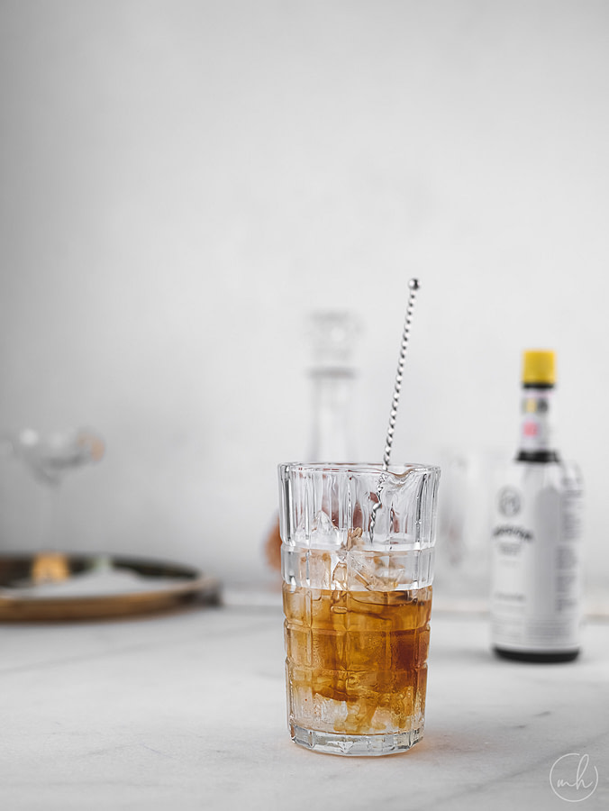 A glass filled with manhattan cocktail with a stirrer, at the background is a bottle of vermouth and whisky