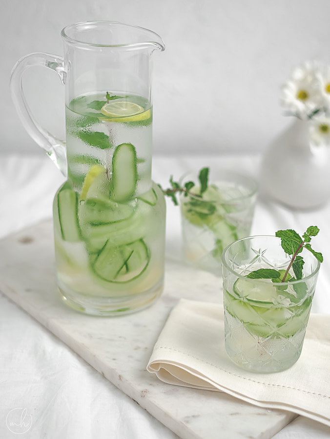Two glasses of cucumber basil lemonade with cucumber slices as garnish. A potted vase with white flowers, cucumber peels, sliced lemon and a half-filled jar of lemonade is in the background.