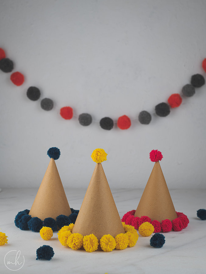 Colourful pompom party hats with red and black party banners in the background and scattered poms