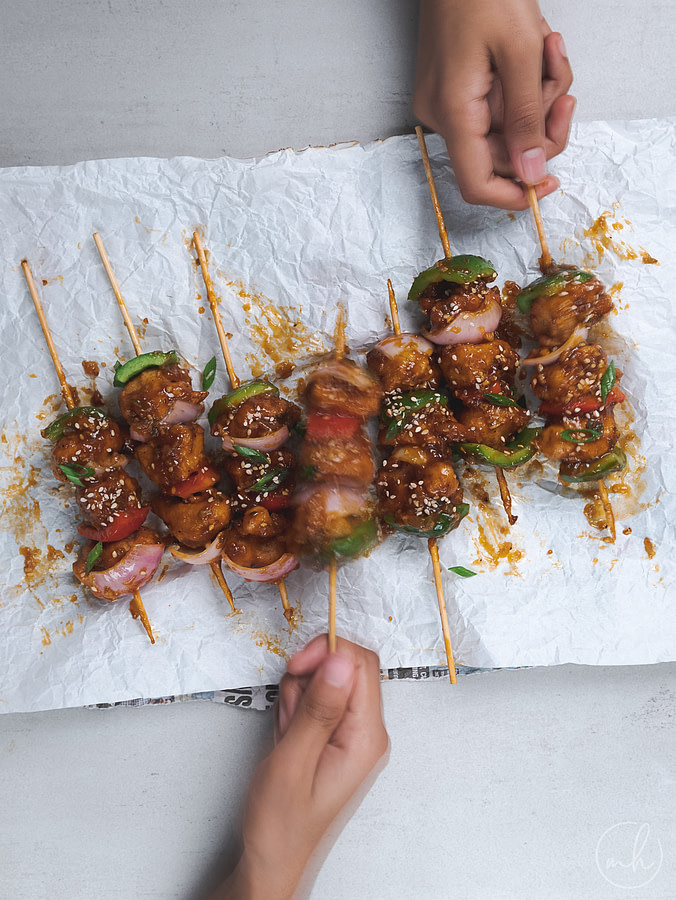 Two hands feasting on cauliflower and paneer manchurian skewers, placed on a white parchment paper.