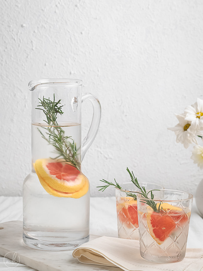 Rosemary grapefruit infused water served in a glass jar and two glasses. It is placed on a marble slate with a cotton cloth. A small pot with white flowers is placed in the background.