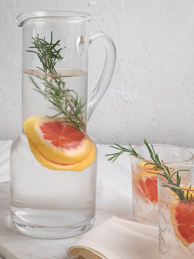 Rosemary grapefruit infused water served in a glass jar and two glasses. It is placed on a marble slate with a cotton cloth. A small pot with white flowers is placed in the background.