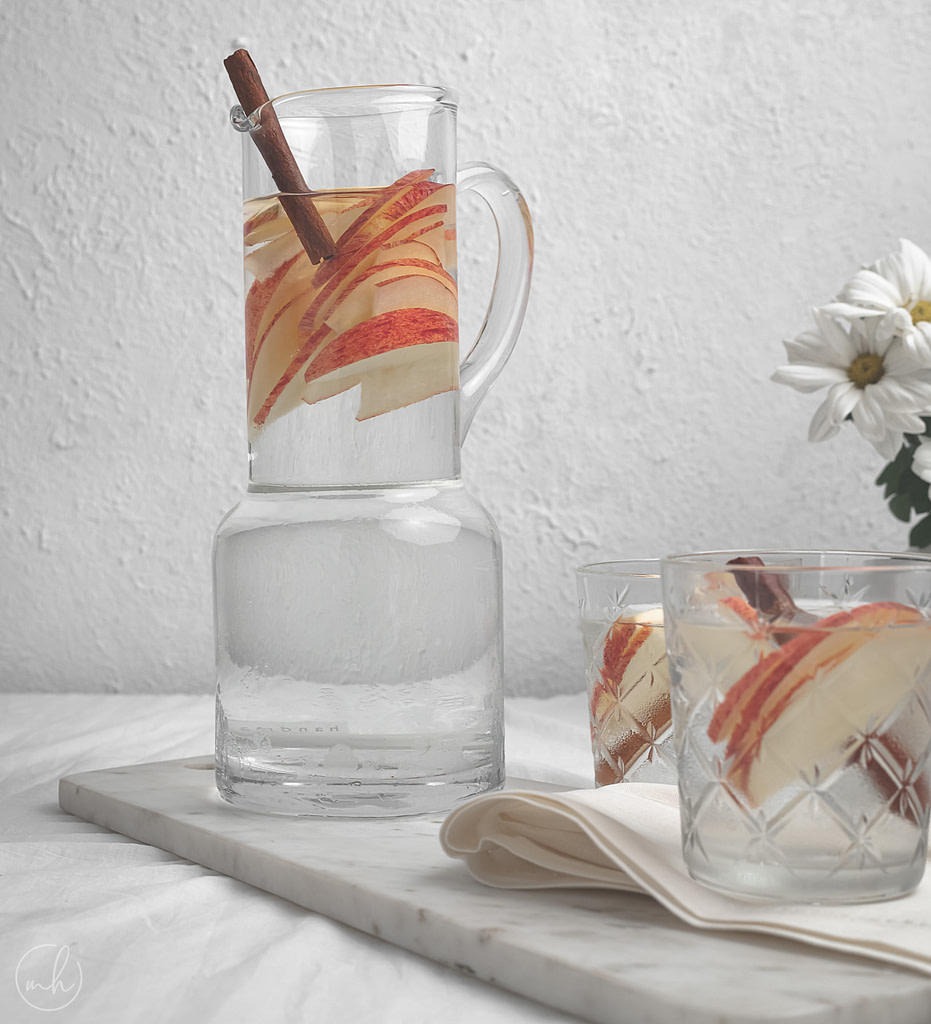 A jar and two glasses filled with apple cinnamon infused water. It's placed on a marble tray with cloth napkin, and a potted vase with white flowers in the background.