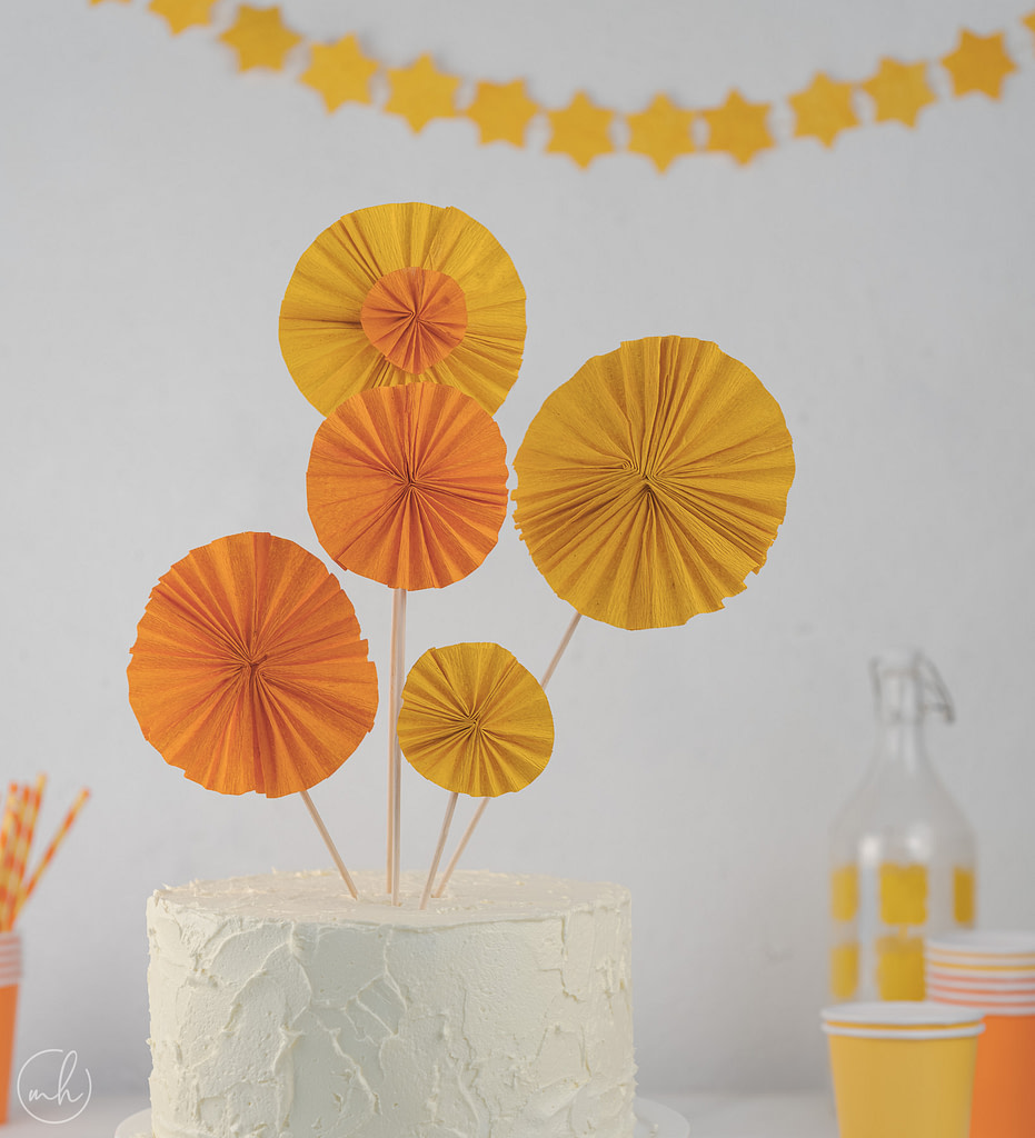 Yellow-orange crepe pinwheel cake topper with yellow party banner bottle and cups in background