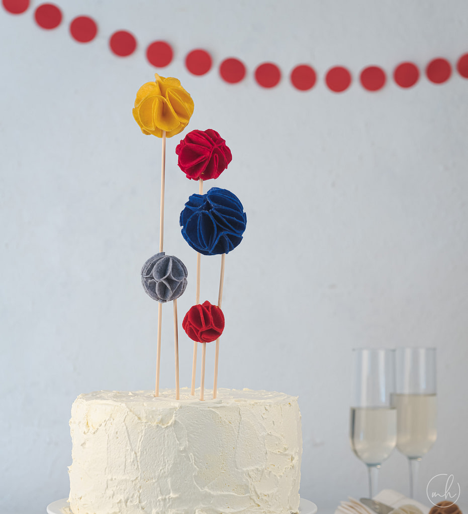 Felt flower cake toppers on white cake champagne glasses plates napkins and red party banners behind