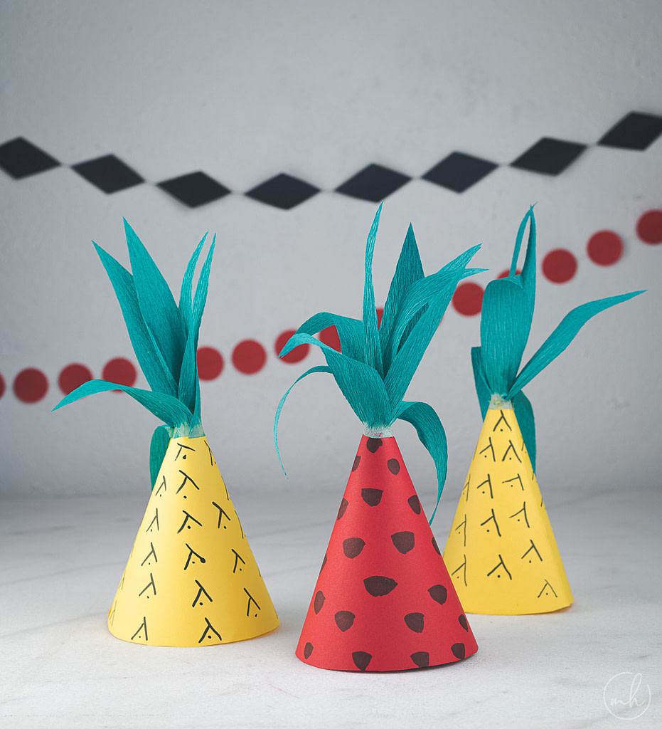 Fruit themed party hats in red, green and yellow with blue and red party banners in the background.