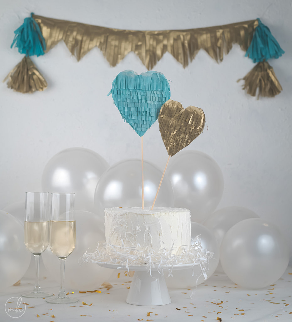 Blue-gold tissue fringe heart cake topper on white cake, blue-gold party banner balloons behind