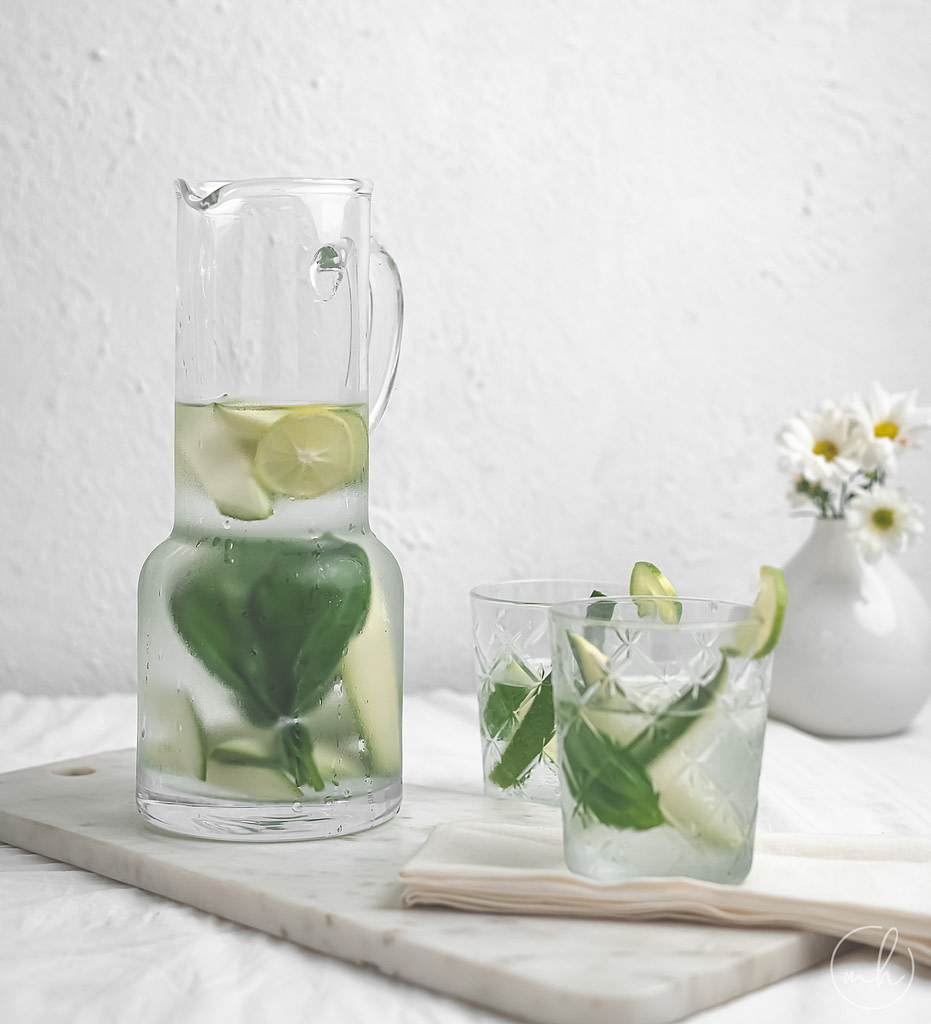Mango basil infused water served in a jar and glass. It is all placed on a marble tile with a cloth napkin. A vase with white flowers is placed in the background.